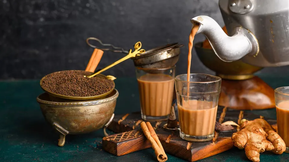 Chai poured from a kettle into a cutting glass, beside a brass bowl of tea leaves, cinnamon sticks and fresh ginger